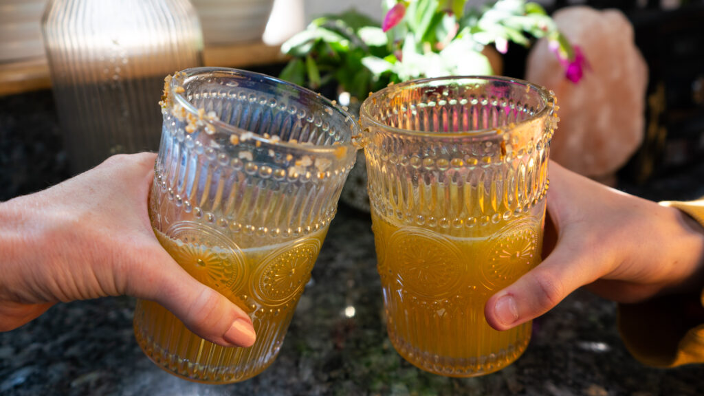 Two people cheersing glasses with yellow juice in it and plants in the background