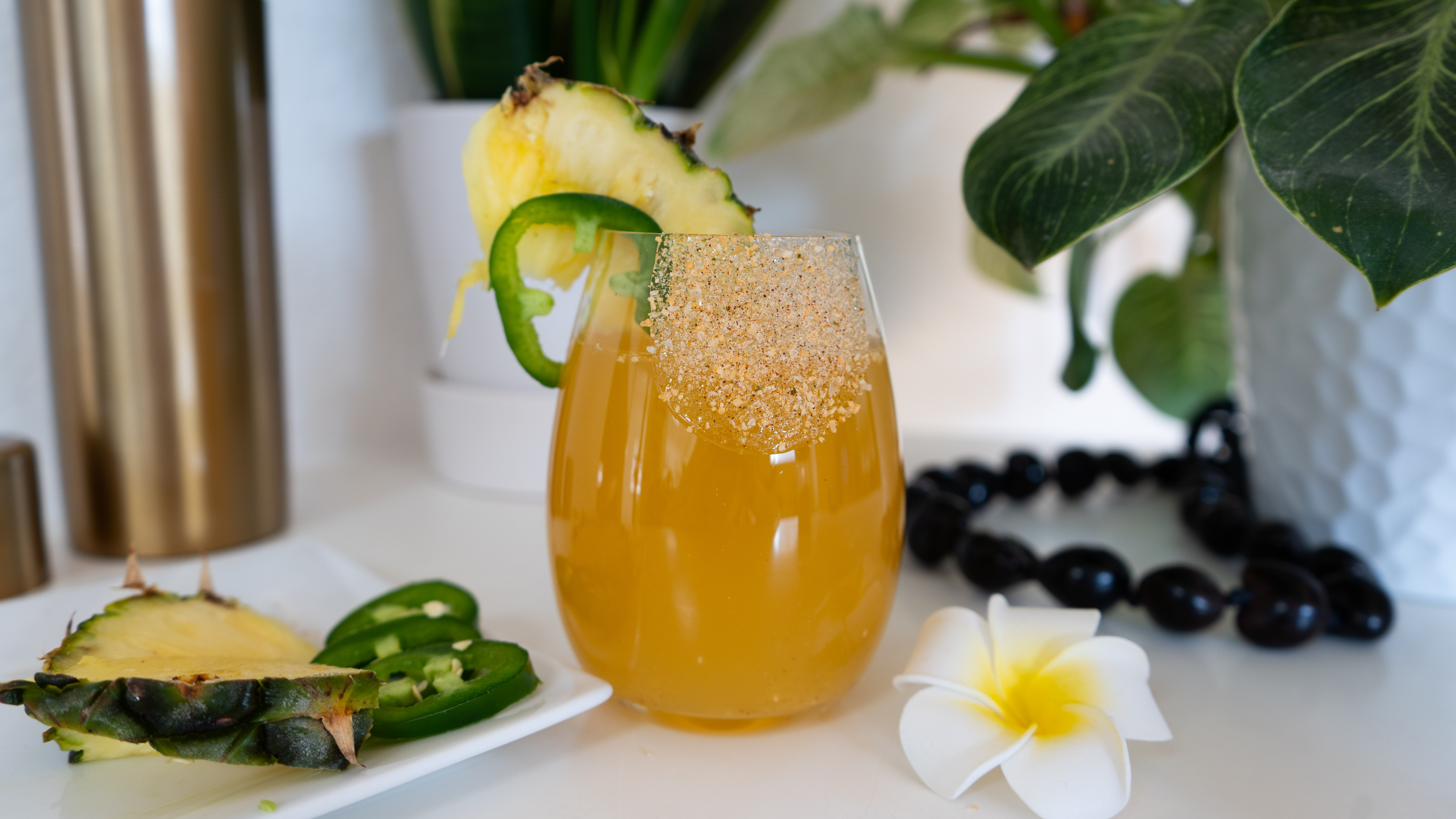 An orange tropical drink with a pineapple, jalapeno and salt on the rim surrounded by plants, flowers and jalapenos