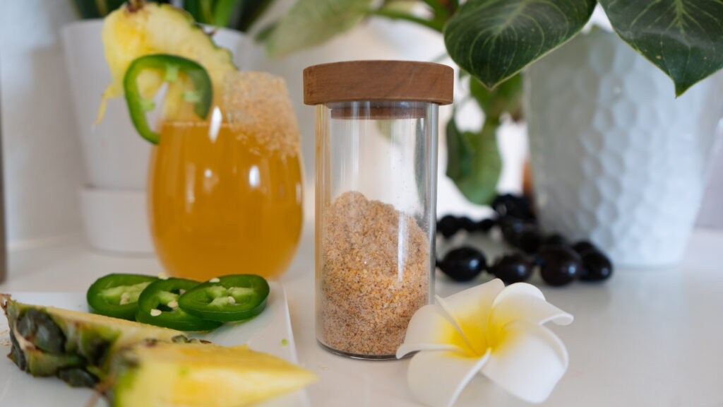 A salt shaker, white and yellow flower, pineapple, jalapeño and tropical drink with plants in the background