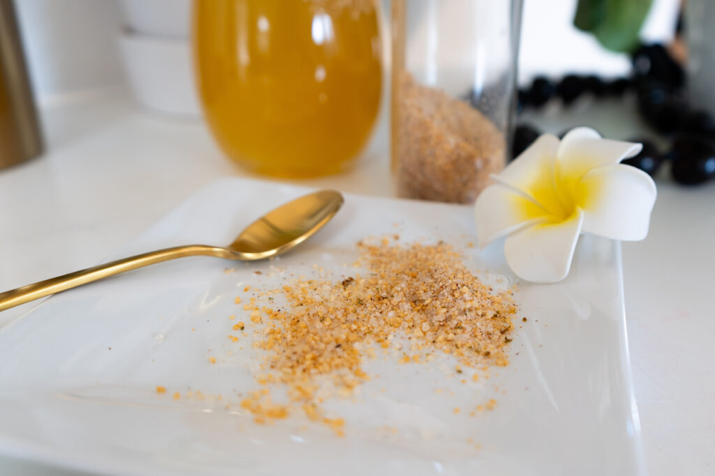 Infused sea salt on a white plate with a gold spoon and white flower, and a drink and salt shaker in the background