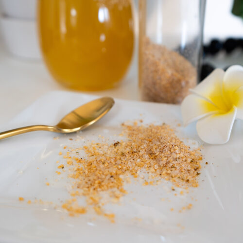 Infused sea salt on a white plate with a gold spoon and white flower, and a drink and salt shaker in the background