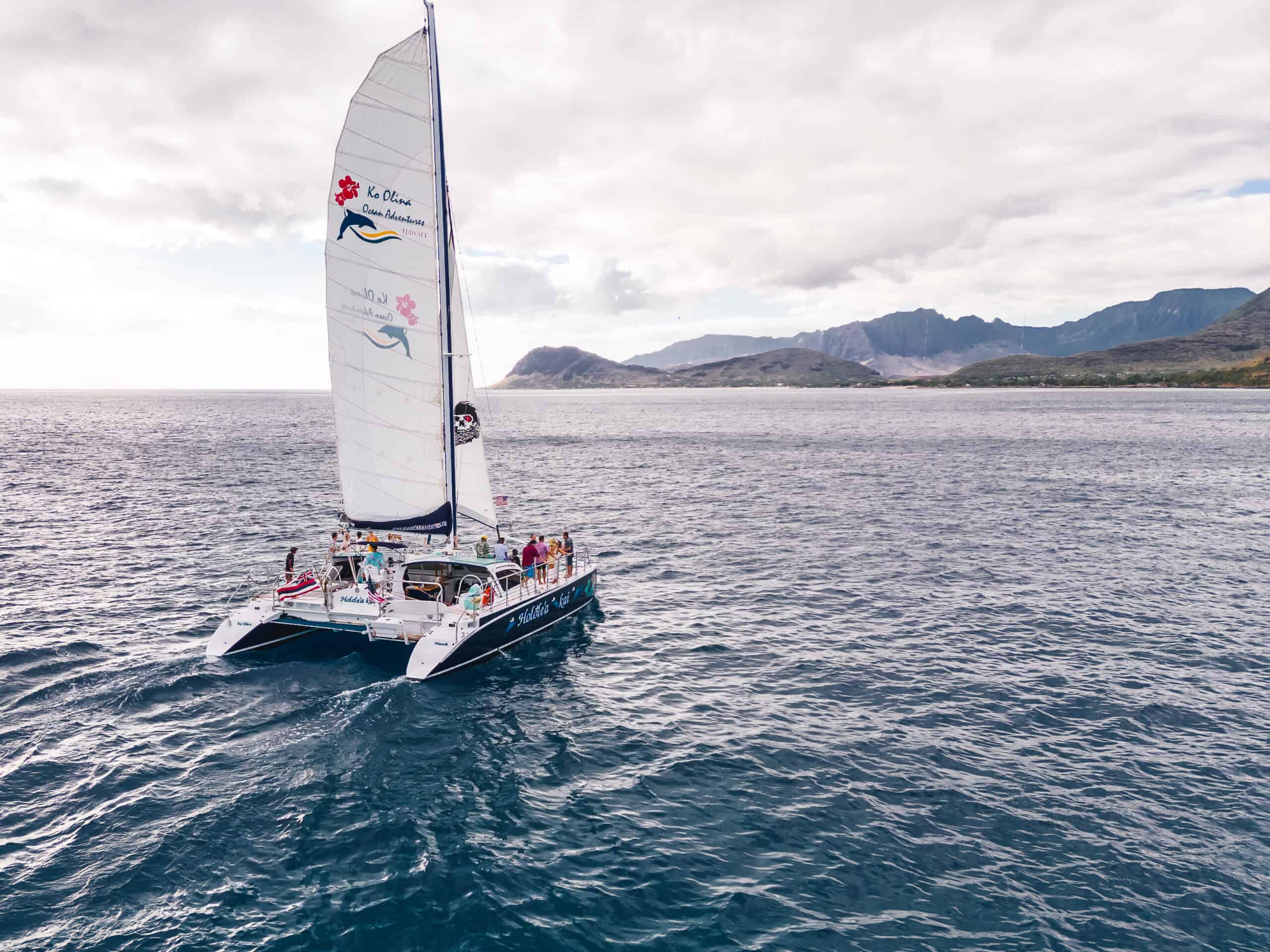 A catamaran sailboat with people on it that says Ko Olina Ocean Adventures on the sail, with an island with rolling hills in the background.