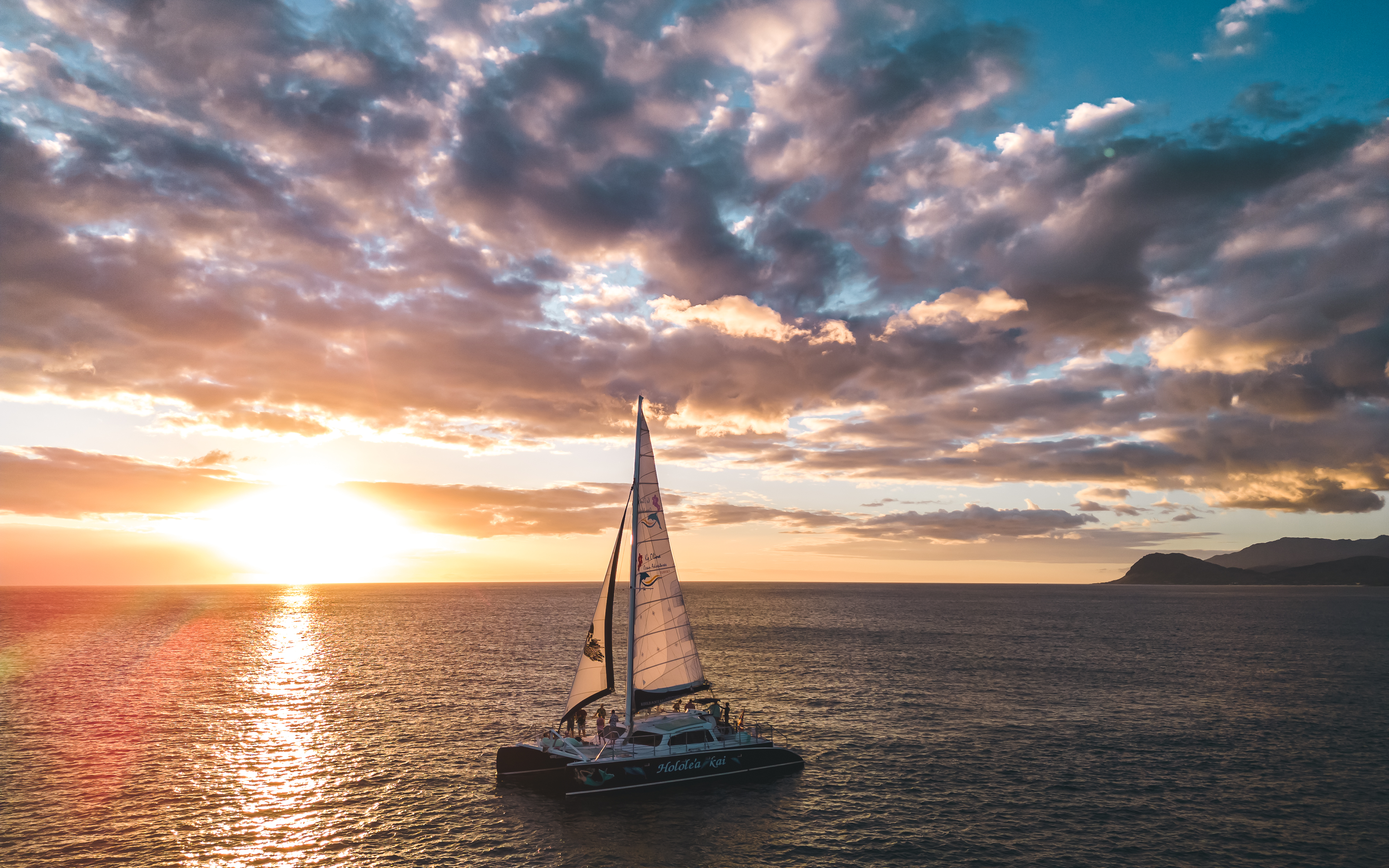 An aerial shot of a sailboat with the sun setting behind it and clouds in the sky