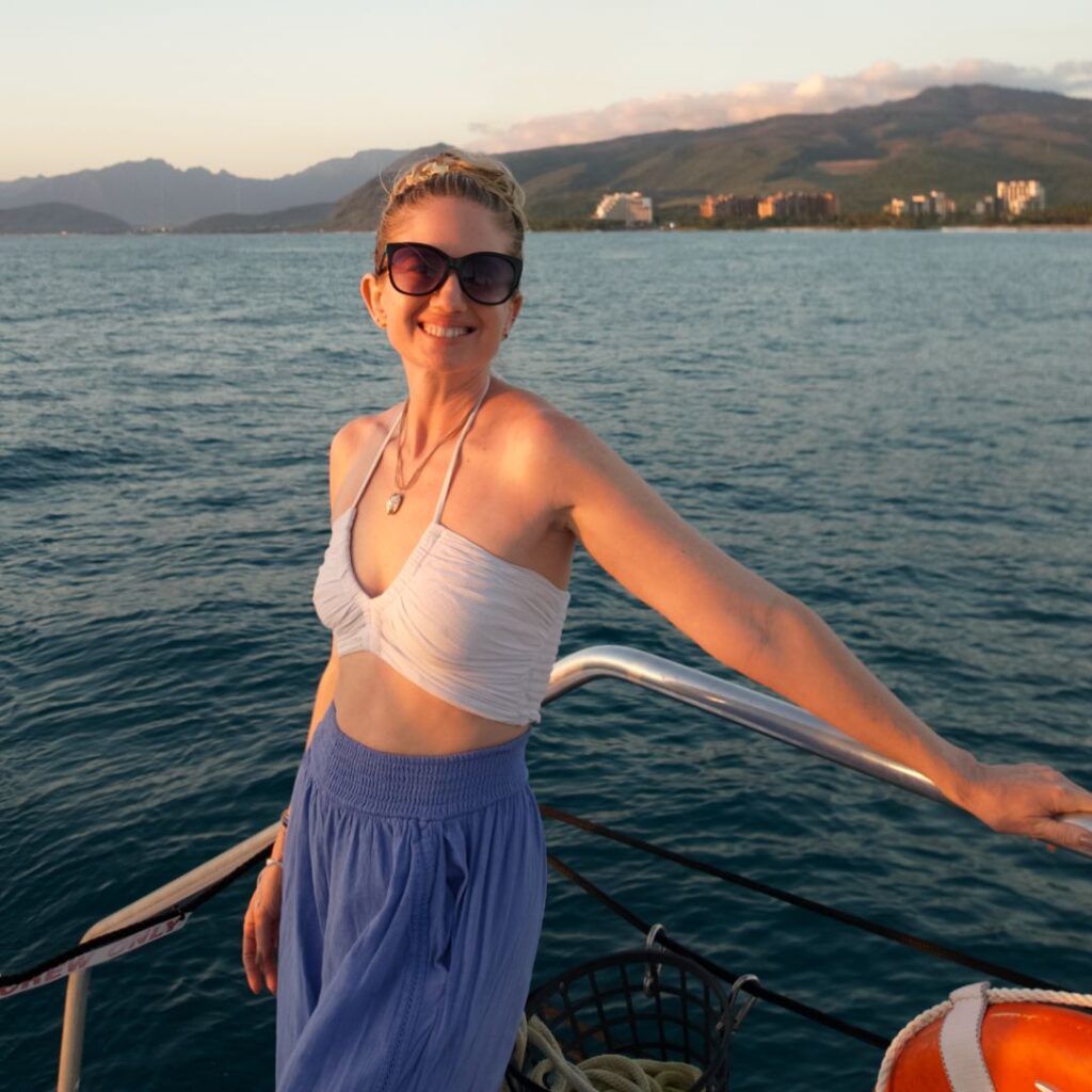 A smiling woman in sunglasses with her hair up, in a white top and blue pants on a boat holding the rail with an island behind her.