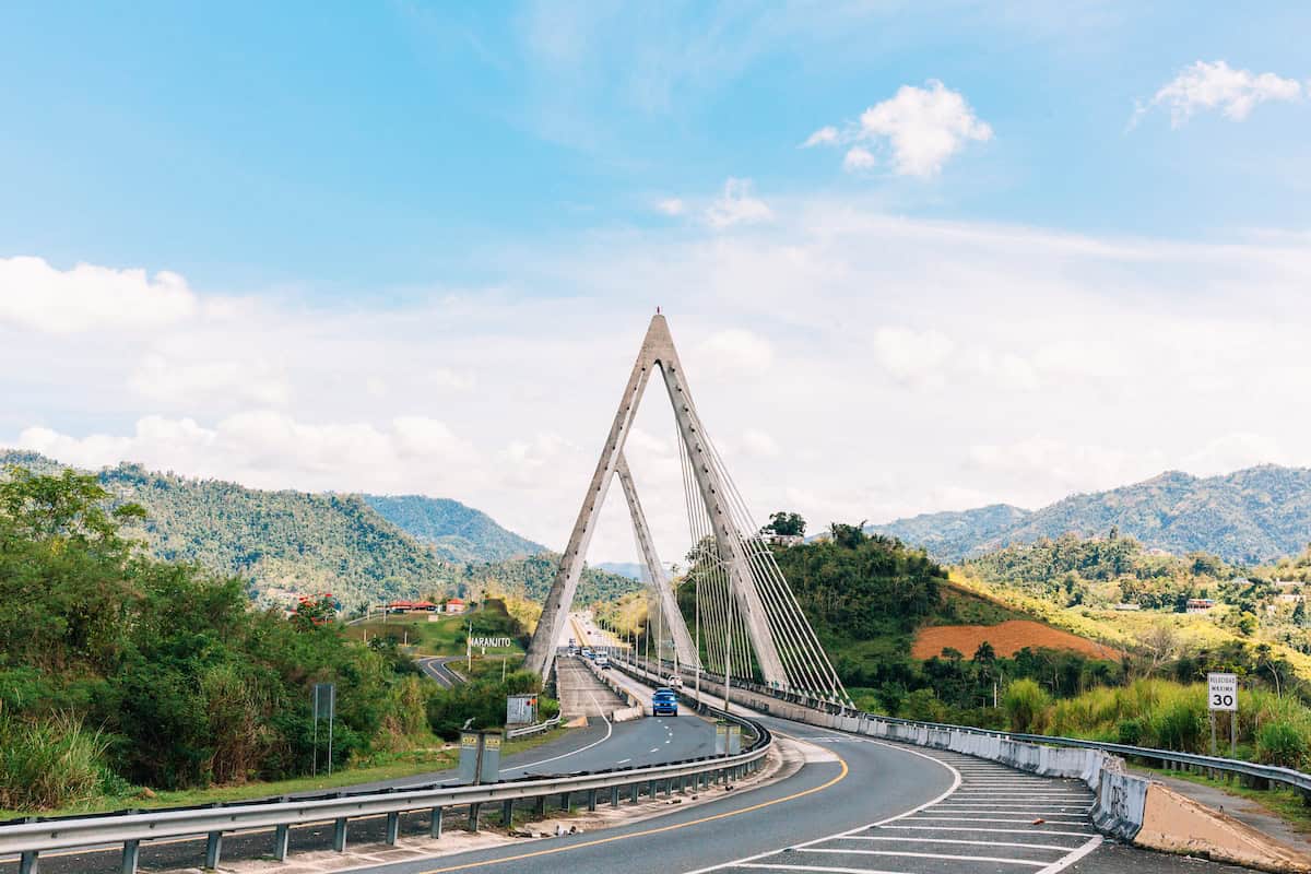 A highway with a white triangular bridge and rolling mountains in the background.