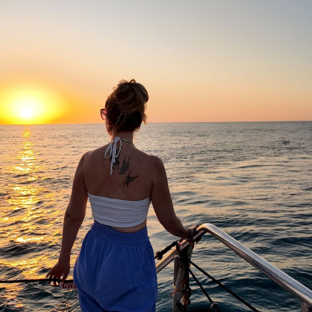 A woman with her hair up, in a white top and blue pants on a boat holding the rail looking at the sunset.