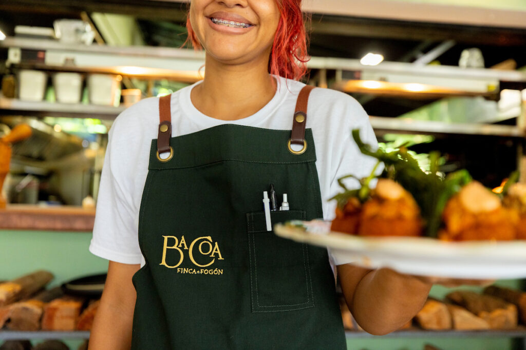 A smiling woman with red hair and a green apron handing a plate of food.